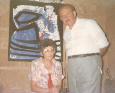 Photo of Mary Rose Caruana, sitting next to Gabriel Caruana on her left hand side, inside The mill - Art, Culture and Crafts Centre, with an abstract painting of Gabriel Caruana as a backdrop.