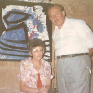 Photo of Mary Rose Caruana, sitting next to Gabriel Caruana on her left hand side, inside The mill - Art, Culture and Crafts Centre, with an abstract painting of Gabriel Caruana as a backdrop.