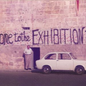 Photo showing the front of the Mill, with a large banner with the words 'Welcome to the Exhibition 1990' written on it. Gabriel Caruana stands below the banner next to the door. A white car is parked adjacent.